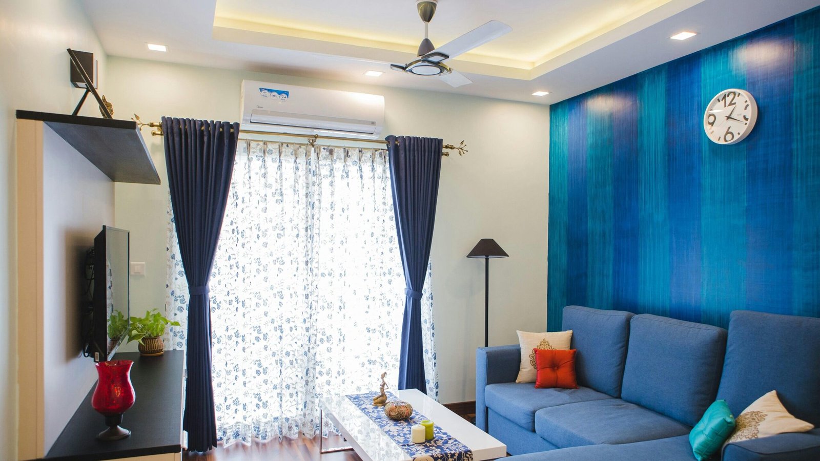Modern living room with blue accents, featuring a sofa, wall clock, and natural light.