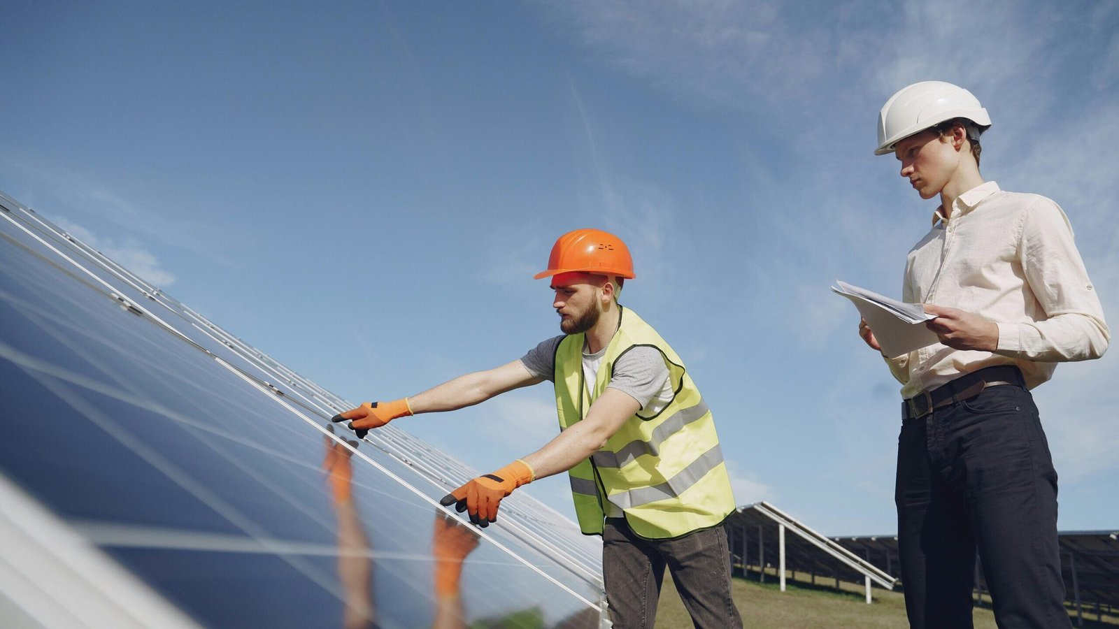 From below of bearded electrical technician in yellow vest and hardhat demonstrating solar panels to young male inspector in formal wear