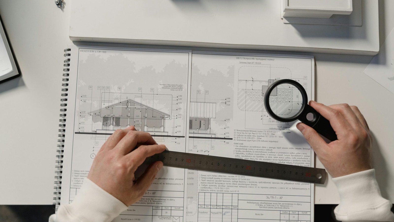 Hands holding a ruler and magnifying glass over architectural floor plans on a white table.